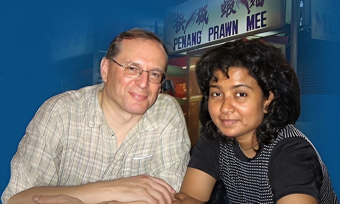 Two people seated together, smiling warmly at the camera, in front of a restaurant sign reading "Penang Prawn Mee." The setting suggests a casual dining experience.