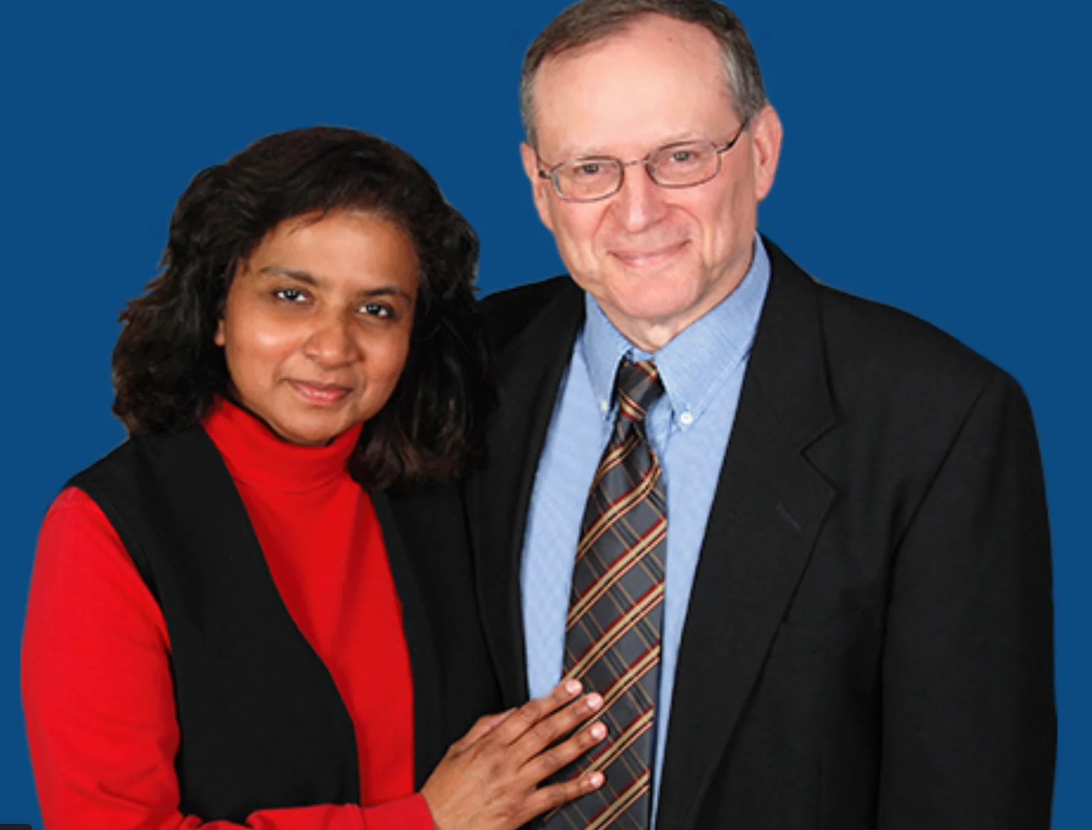 A woman in a red turtleneck and a man in a suit smile warmly against a blue background, showcasing a professional yet friendly atmosphere.