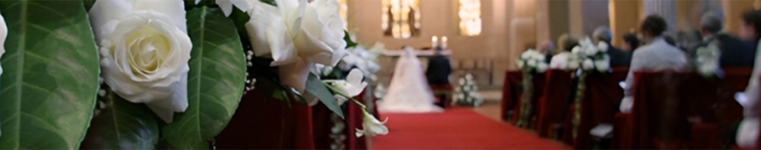 White roses and greenery adorn the aisle, leading to a wedding ceremony at a beautifully decorated venue, with guests seated in the background.