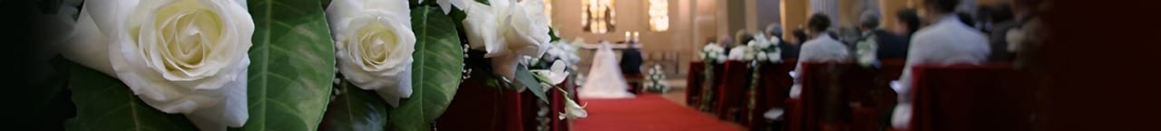 White roses and greenery adorn the aisle, leading to a wedding ceremony at a beautifully decorated venue, with guests seated in the background.