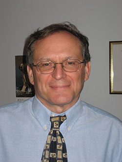 Smiling middle-aged man with glasses, wearing a light blue shirt and patterned tie, against a neutral background with framed art.