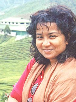Smiling woman with curly hair in a scenic tea plantation. The lush green landscape and distant buildings create a serene, natural backdrop.